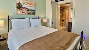 A neatly made bed with white linens and a brown textured blanket, a bedside lamp, and a painting on the wall.