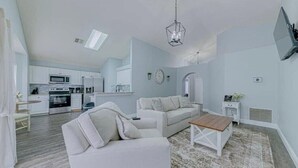 Una sala de estar moderna con un sofá blanco, una mesa de centro de madera y una cocina al fondo.