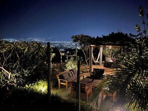 An outdoor patio with a wooden table and chairs, a hammock, and a view of a cityscape at night.