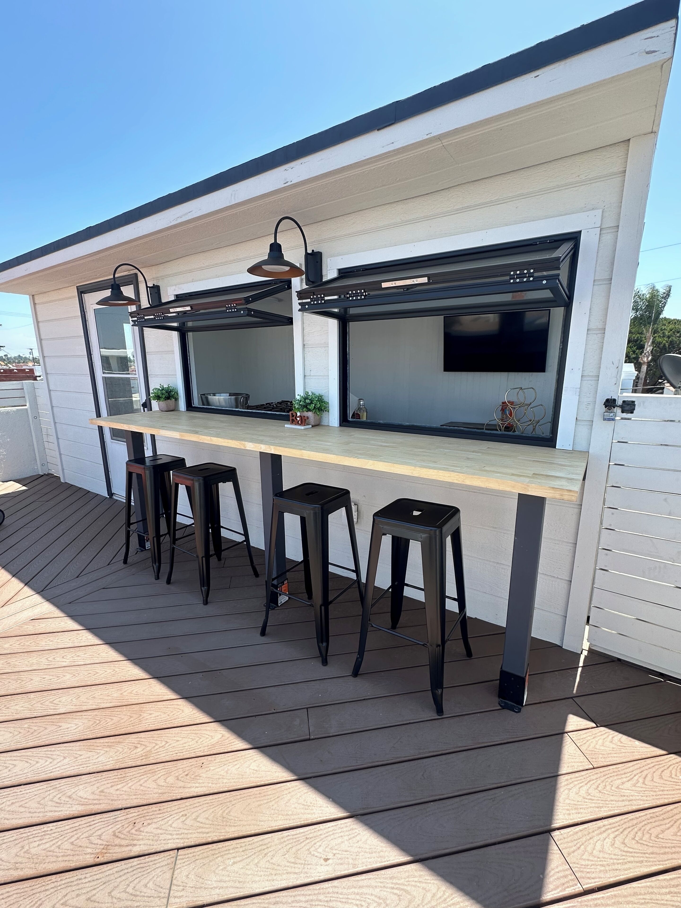 Un espace cuisine extérieur doté d’un plan de travail en bois, de tabourets noirs et d’un luminaire noir suspendu au plafond.