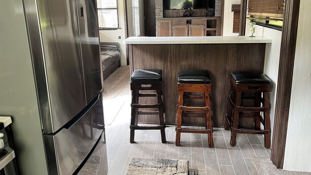 A modern kitchen with a built-in oven, a large TV, and wooden bar stools. - Modern RV come “GLAMP” in style. (Grand Rapids)