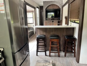 A kitchen with a built-in TV, wooden bar stools, and a stainless steel refrigerator. - Modern RV come “GLAMP” in style. (Grand Rapids)