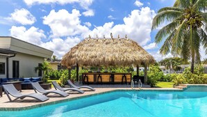 Une piscine avec des chaises longues, un bar à toit de chaume et un palmier.