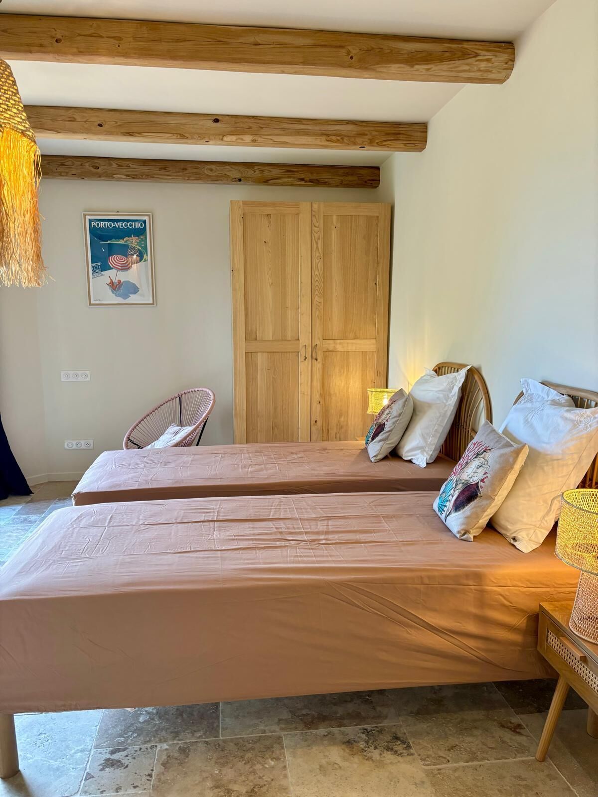 Une chambre avec deux lits, des poutres en bois, un fauteuil en osier, une armoire en bois et une affiche sur le thème de la plage.