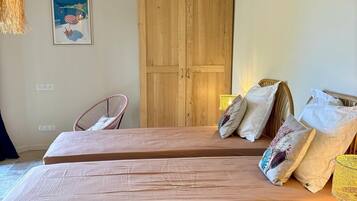 Une chambre avec deux lits, des poutres en bois, un fauteuil en osier, une armoire en bois et une affiche sur le thème de la plage.