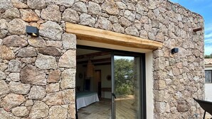 Eine Fassade aus Stein mit einer Holzdielenveranda und einer Schiebetür zu einem Innenraum mit Bett und Pendelleuchte.