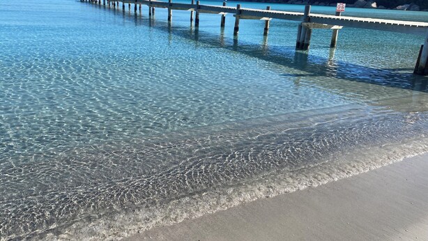Eine Pier, die sich über klares, blaues Wasser zu einem Sandstrand mit einer gebirgigen Kulisse erstreckt.