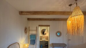 Ein Schlafzimmer mit einem großen Bett, Holzbalken, einer Hängelampe, einem Spiegel und Blick ins Bad.