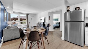 A modern kitchen with a stainless steel refrigerator, a dining area with a round table and chairs, and a living area with a sofa and a TV.