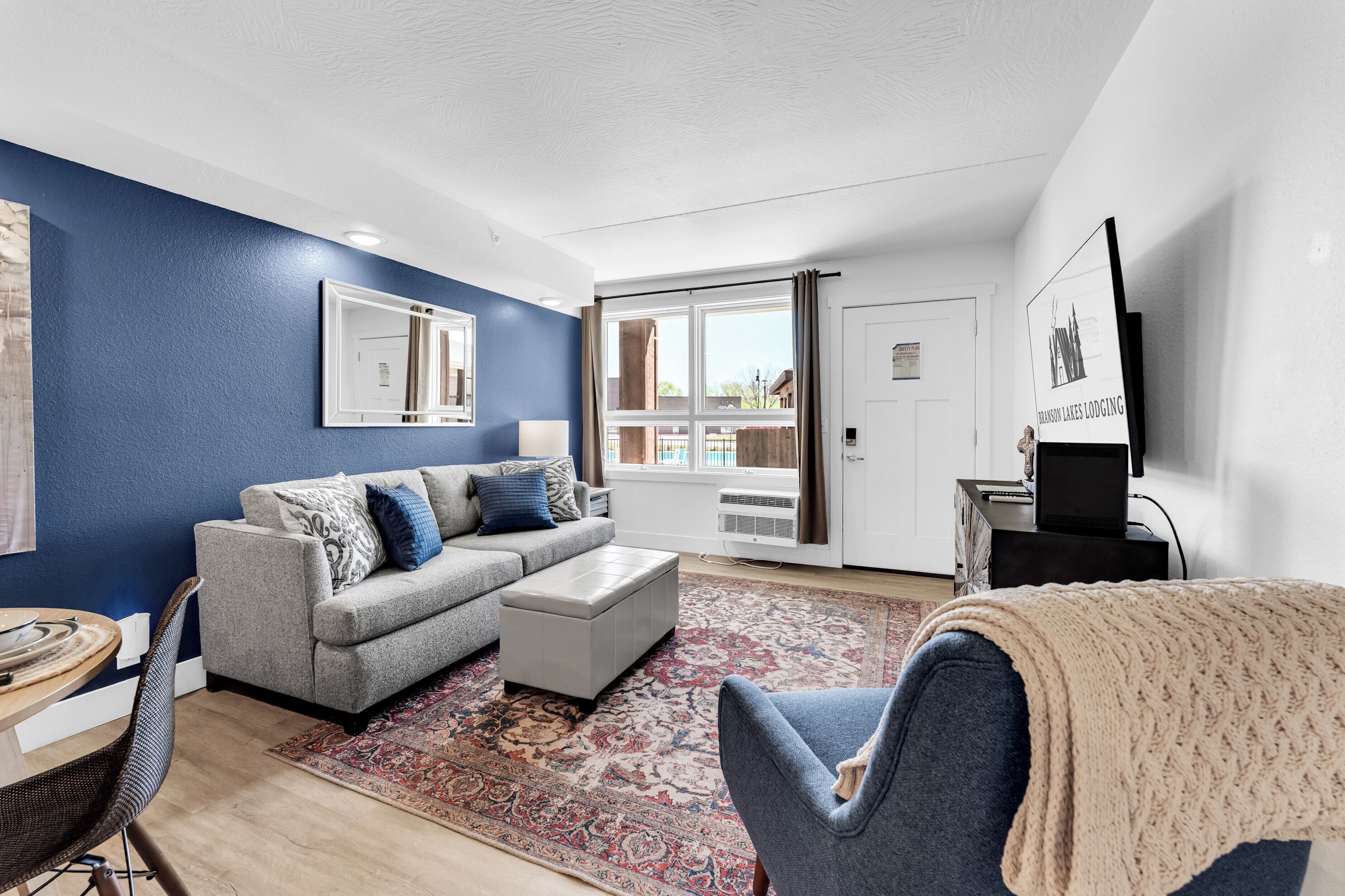 A modern living room with a grey sofa, a patterned rug, and a large window with curtains.