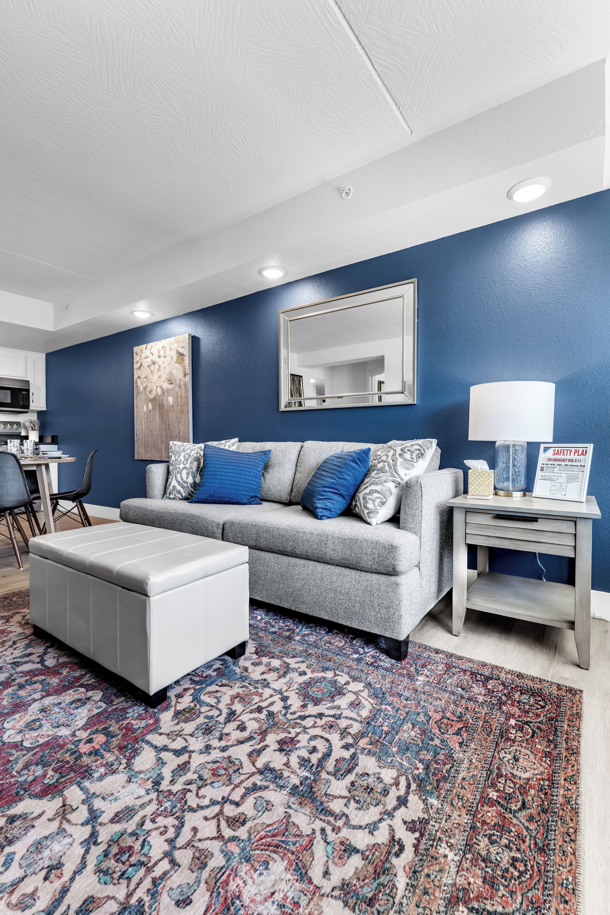 A living room with a grey sofa, patterned rug, and a small side table with a lamp and a safety plan sign.