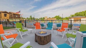 An outdoor seating area with colorful chairs and a central fire pit, overlooking a swimming pool and a playground.
