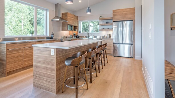 Une cuisine moderne dotée d’un grand îlot central, de tabourets de bar, de meubles en bois, d’appareils électroménagers en acier inoxydable et de luminaires suspendus.