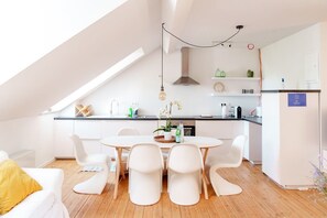 A modern kitchen with white cabinetry, a round dining table, and white chairs. There is a skylight above the kitchen, and a hanging light fixture. - Duplex Apartment - Midi Station / Nespresso / kitchen (Saint-Gilles)