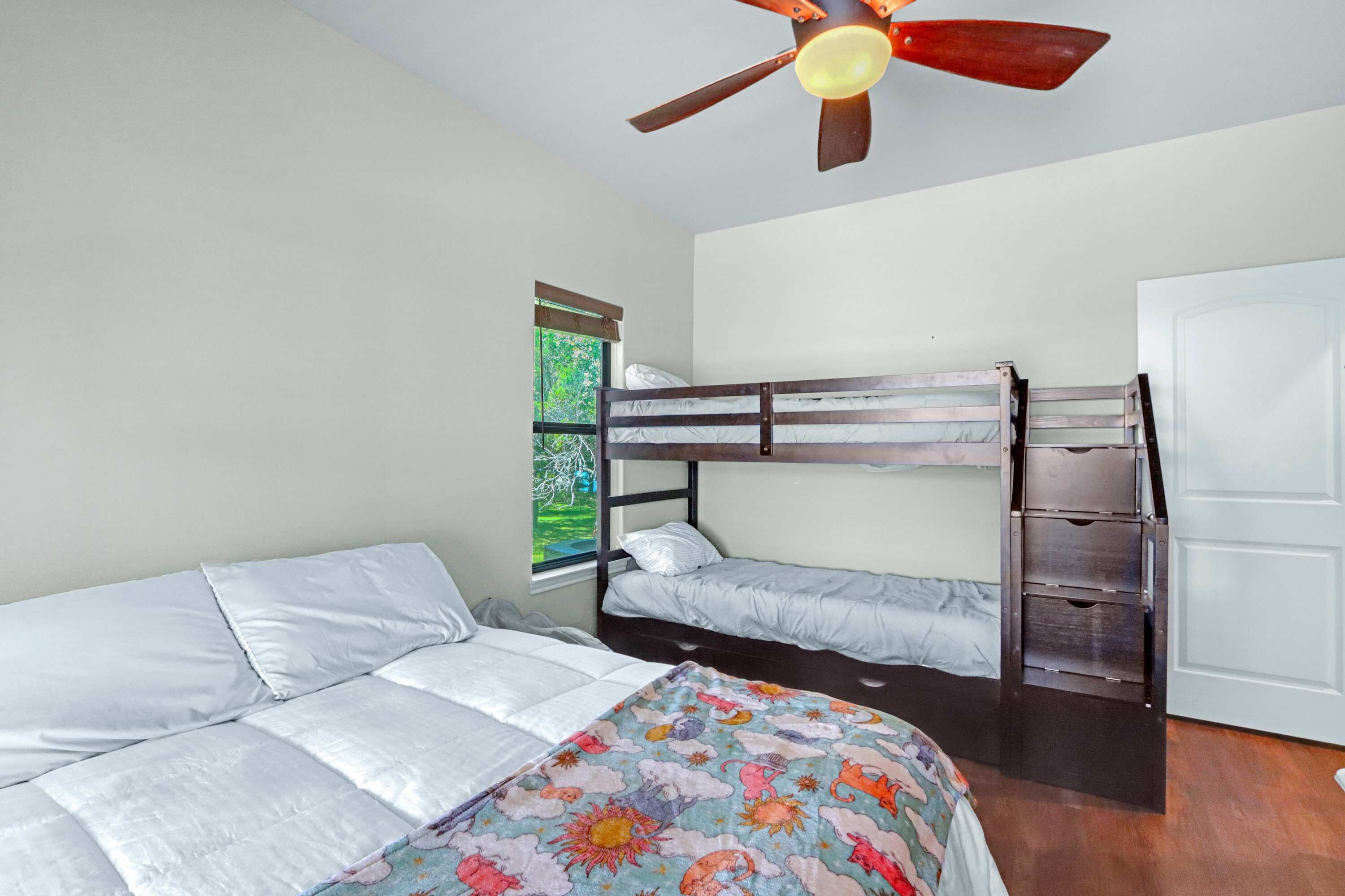 A bedroom with a bunk bed, a single bed with a floral bedspread, a ceiling fan, and a window with a view of greenery.