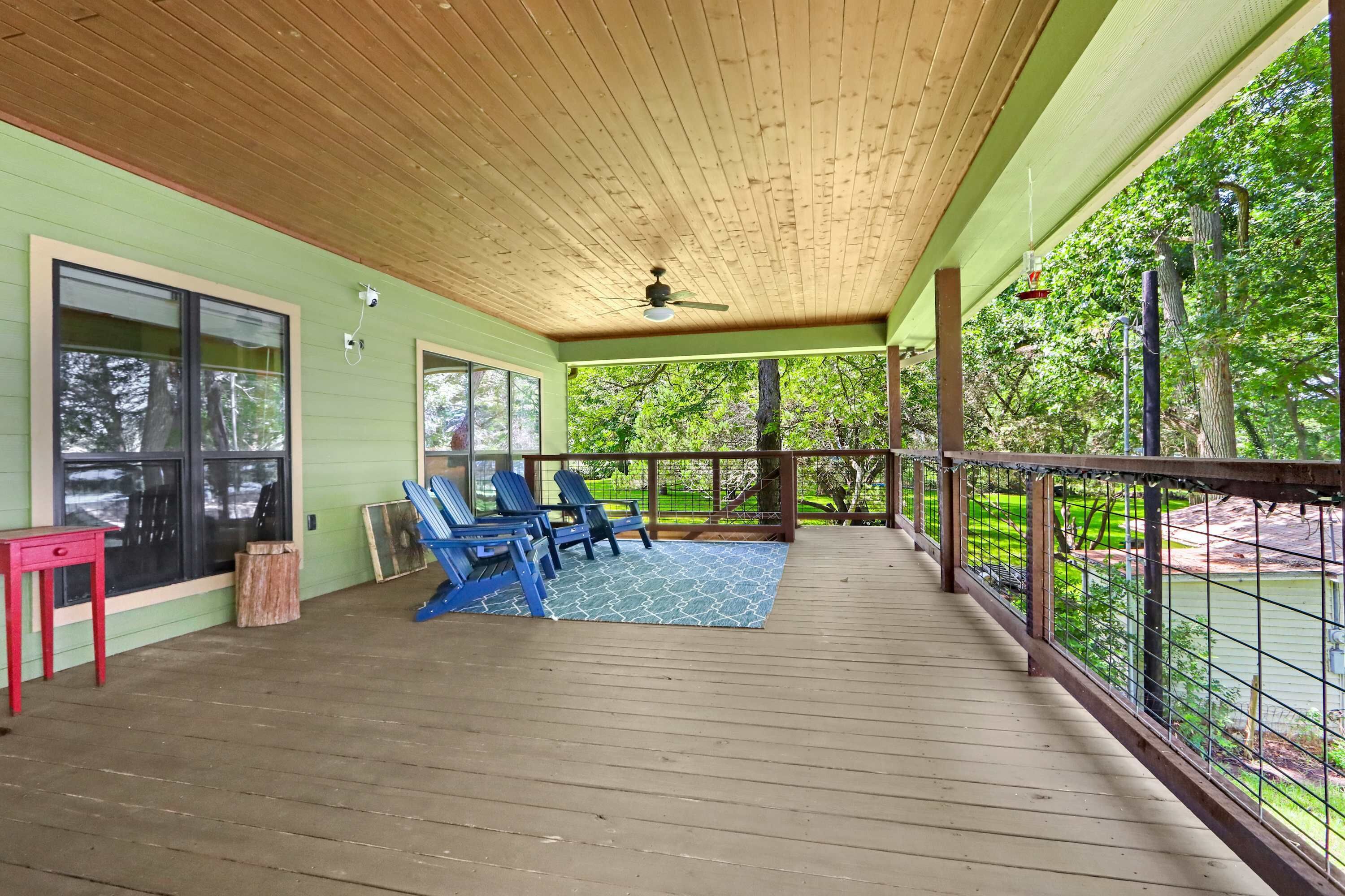A covered outdoor area with a wooden deck, blue chairs, a red table, and a view of a green landscape.