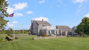 A two-story house with a stone foundation, a front porch, and a flagpole with an American flag. - Coastal Farmhouse Retreat in Little Compton, RI – Your Perfect Seaside Getaway (Little Compton)