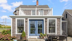 A two-story house with a blue front door, a covered patio with a table and chairs, and a small garden with various plants. - Coastal Farmhouse Retreat in Little Compton, RI – Your Perfect Seaside Getaway (Little Compton)