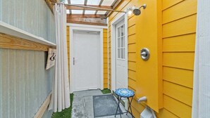 Un estrecho pasillo exterior con techo de madera, pared amarilla y una pequeña mesa azul.