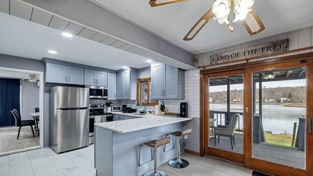 A modern kitchen with a large island, stainless steel appliances, and a view of a lake through the sliding glass doors. - Direct Lakefront House in Poconos (Tobyhanna)