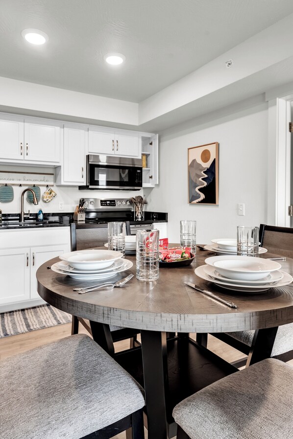 Una cocina moderna con armarios blancos, un microondas y una mesa de comedor preparada para cuatro personas, con platos y vasos.