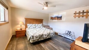 Un dormitorio con una cama de madera, una mesita de noche con lámpara, un ventilador de techo, una silla, un televisor y un perchero en la pared.