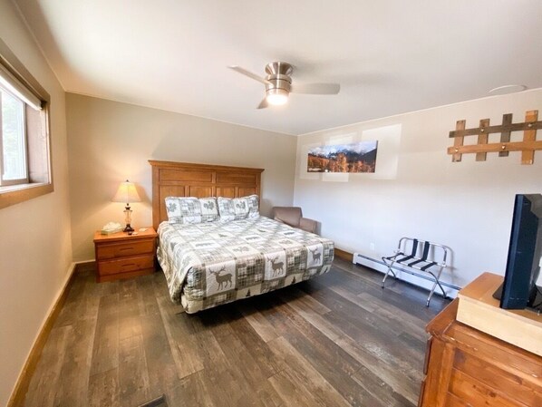 A bedroom with a wooden bed, a nightstand with a lamp, a ceiling fan, a chair, a TV, and a wall-mounted rack. - Relaxing Getaway in our King Kitchenette at Lodges of Oak Creek! (Oak Creek)