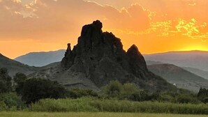 A natural landscape with a prominent rock formation in the foreground and a sunset in the background.