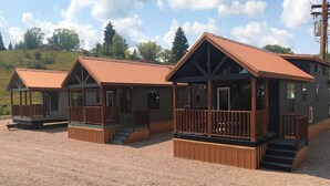 A row of cabins with wooden decks and metal roofs, set on a gravel ground with a hilly background.