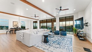 A spacious living room with a large sectional sofa, a coffee table, and a blue patterned rug. There are two ceiling fans, a TV mounted on the wall, and a dining area with a table and chairs. - Luxury LBJ Lakefront w/sunset views, pool, hot tub, game room/pingpong; perfect for large gatherings! (Granite Shoals)
