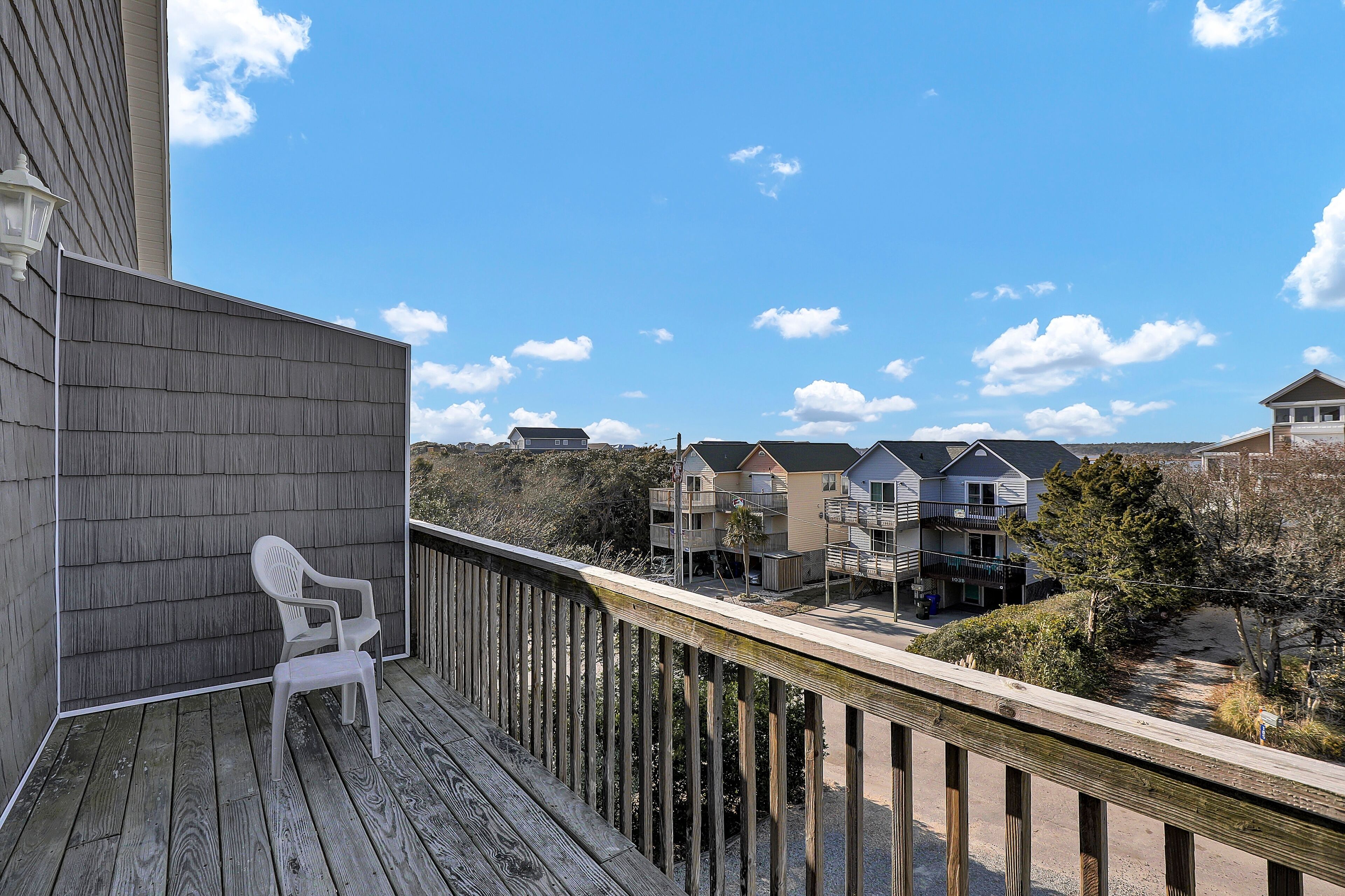 A wooden deck with a white chair, overlooking a row of multi-story buildings with balconies and a clear blue sky.