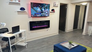 A modern living room with a flat-screen TV, a fireplace, a desk with a laptop, and a blue ottoman. - King Suite | 1mi to The Battery + Mins to Midtown! (Atlanta)