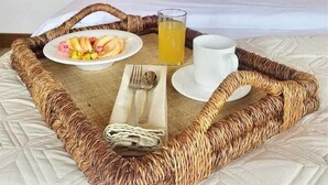 Un lit bien fait, avec une tête de lit ornée de motifs de feuilles, une table de chevet sur laquelle est posé un vase de fleurs, et un plateau de petit-déjeuner contenant des fruits, un verre de jus et une tasse, le tout placé sur un tapis tissé.