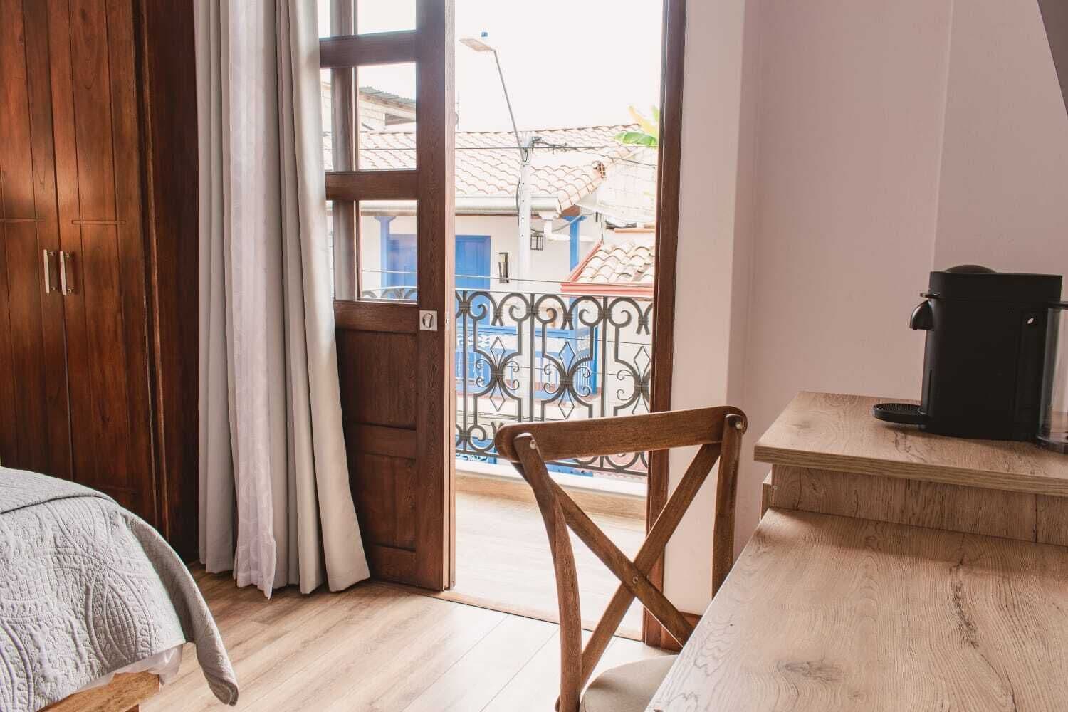 A room with a wooden wardrobe, a bed with a white blanket, a wooden chair, a small table with a coffee maker, and a balcony with a metal railing.