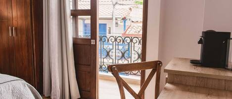 A room with a wooden wardrobe, a bed with a white blanket, a wooden chair, a small table with a coffee maker, and a balcony with a metal railing.