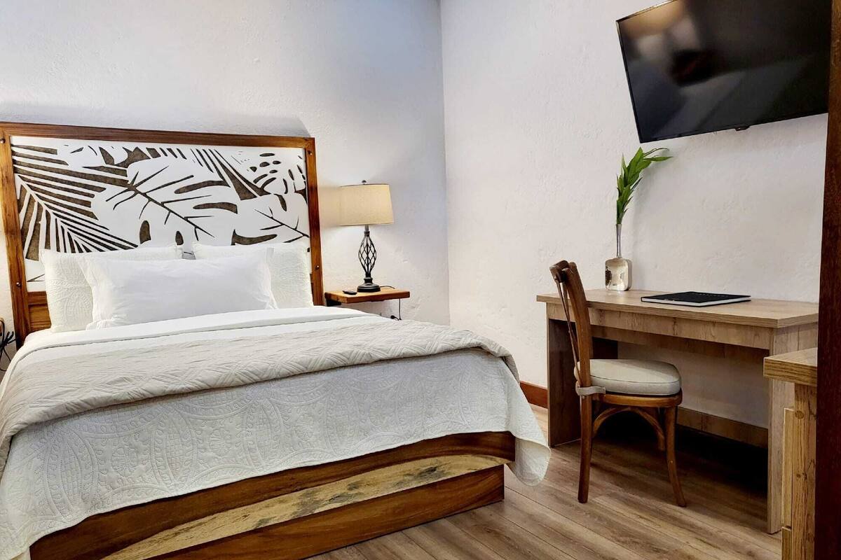 A hotel room with a wooden bed, a desk, a chair, a TV, and a plant.