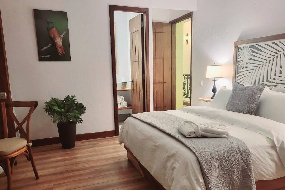 A bedroom with a wooden bed, a chair, a potted plant, and a painting on the wall.