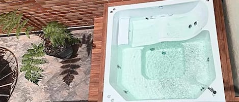 A hot tub with a rectangular shape and a wooden border, placed on a patio with a brick wall and potted plants.