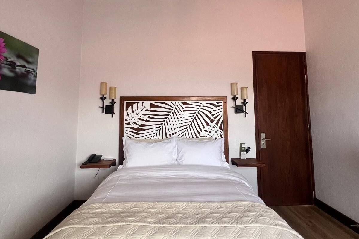 A bedroom with a bed, wooden ceiling beams, a decorative headboard, bedside lamps, and a wooden door.