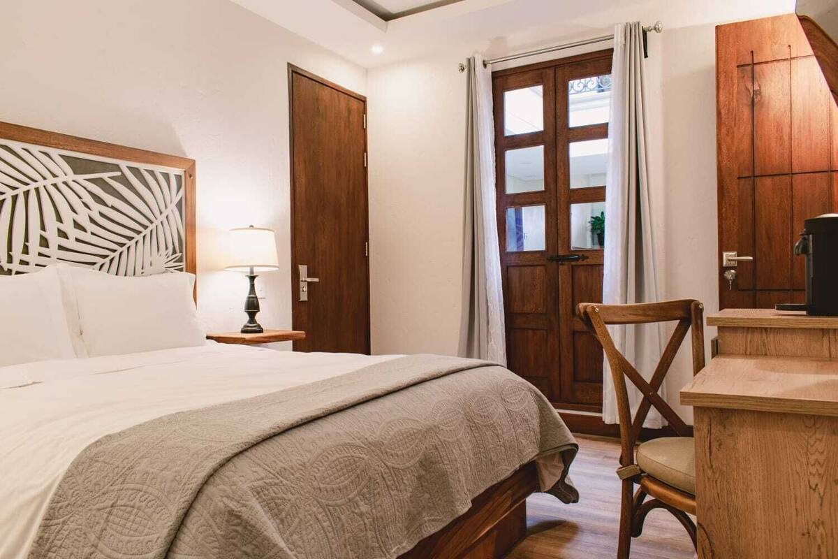 A bedroom with a bed, a nightstand, a chair, a wooden desk, a coffee machine, and a door with a window.