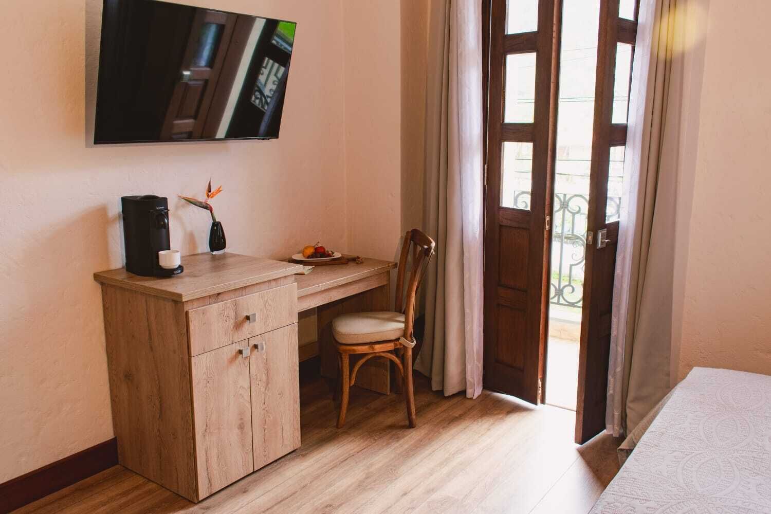 A hotel room with a wooden desk, a chair, a coffee maker, a plate of fruit, and a TV mounted on the wall.