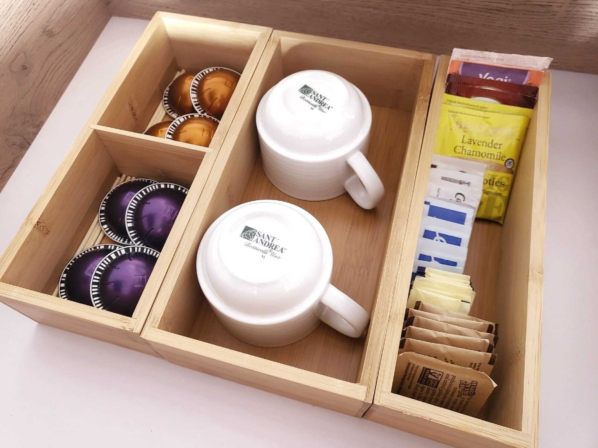 A wooden box containing two white coffee mugs, several purple and brown coffee cups, and packets of coffee grounds.