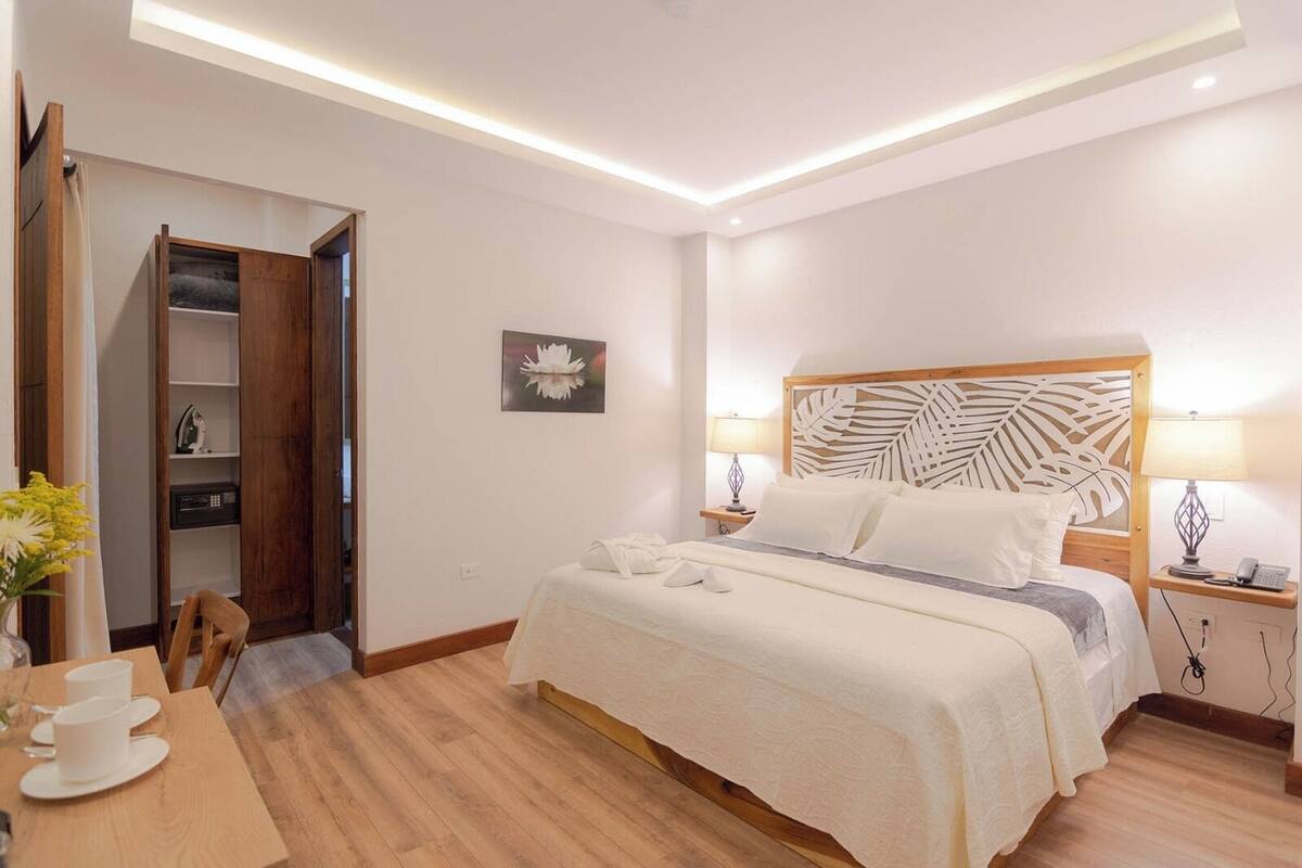 A hotel room with a bed, bedside tables, a desk with cups, a chair, a wardrobe, and a picture on the wall.