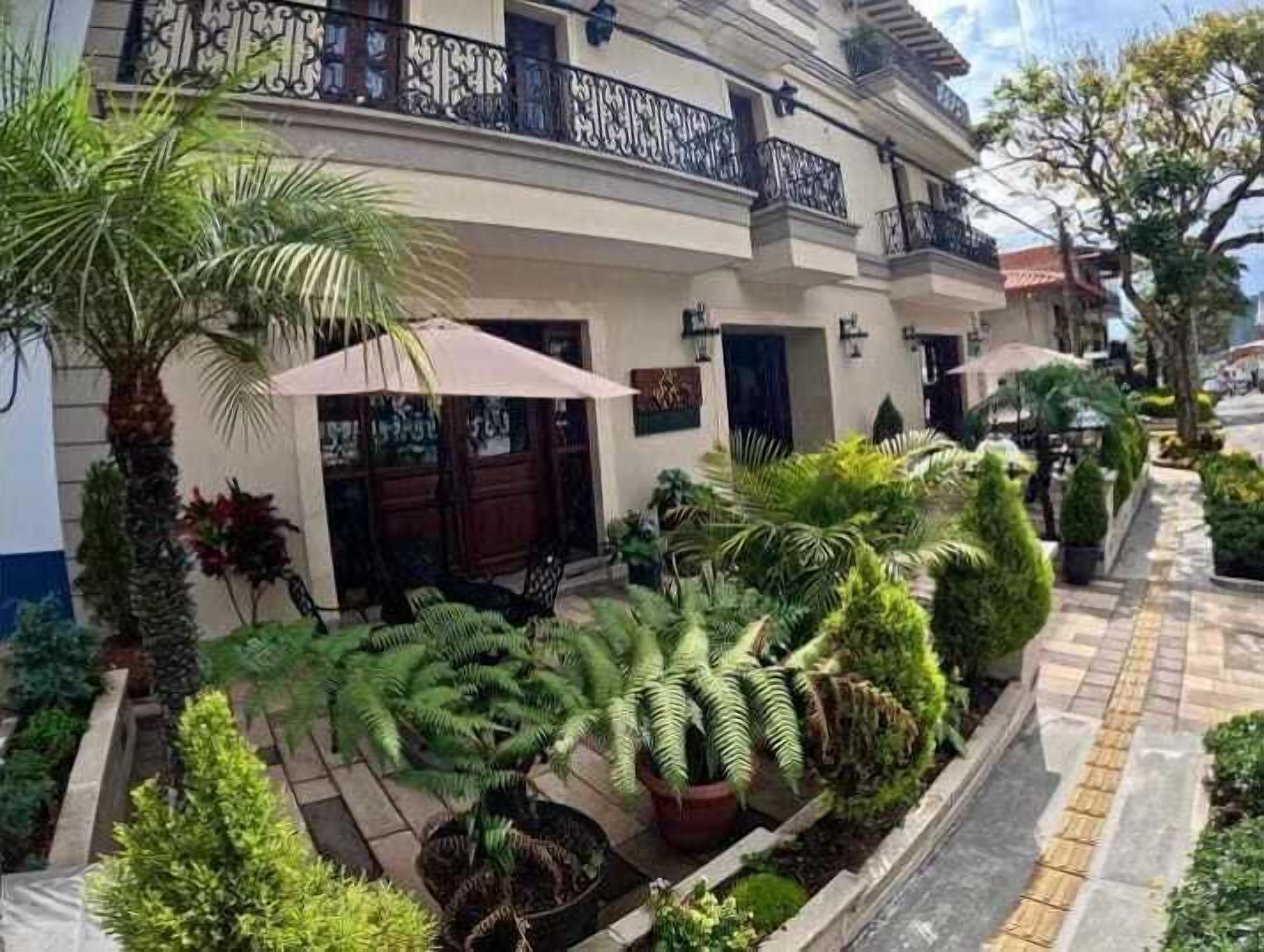 Un bâtiment doté d’une façade incurvée, d’un balcon orné de balustrades noires et d’un jardin bien entretenu, agrémenté de diverses plantes et d’un espace patio.