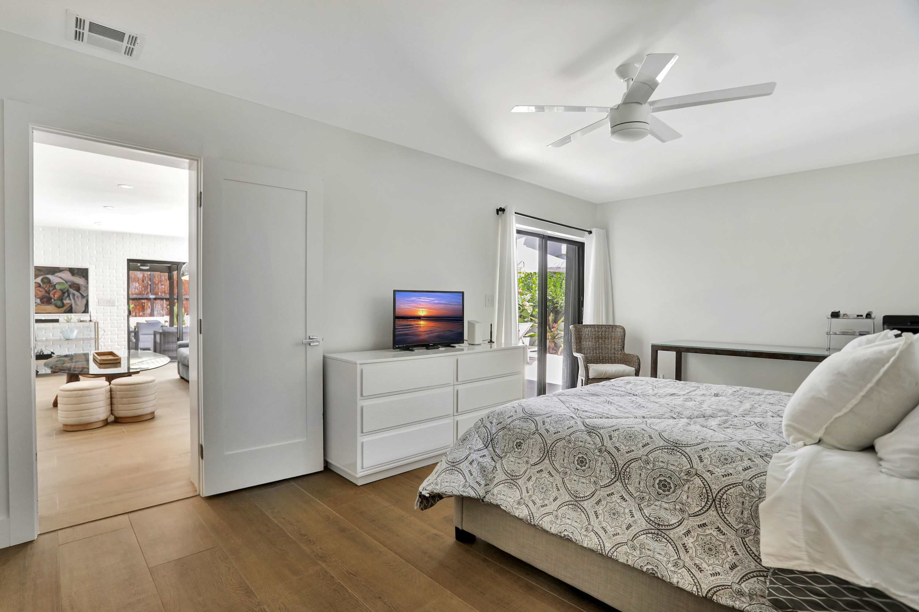 A bedroom with a bed, a TV, a ceiling fan, and a view of the outside.