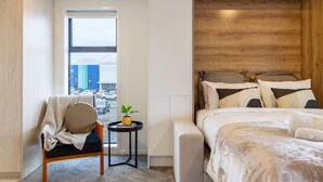 A modern hotel room with a large bed, a seating area with a chair and a small table, and a view of the street through a large window. - Luxurious Studio: Prime location, City Centre (Christchurch)