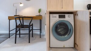 A compact room with a washing machine, a desk with a chair, a lamp, and a small plant. - Single Level Studio, CBD Escape: King bed (Christchurch)