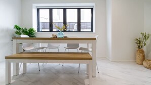 Een moderne eetruimte met een houten tafel en witte stoelen, een raam met zonwering en potplanten.