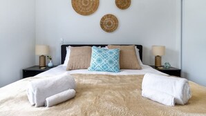 Een net opgemaakt bed met een zachte, beige deken, twee witte handdoeken en decoratieve kussens. Daarboven hangen drie geweven wanddecoraties.