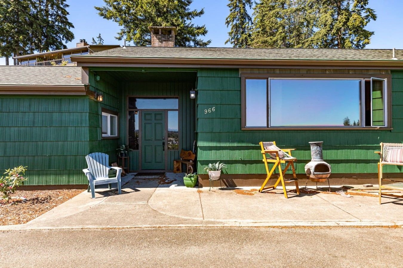 A green house with a number '966' on the door, a front porch with a blue chair, a yellow chair, and a small table.